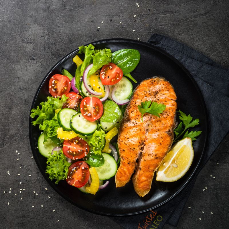 Image of black plate with bright orange salmon filet and a colorful salad on a gray napkin with Twisted Paleo on the napkin